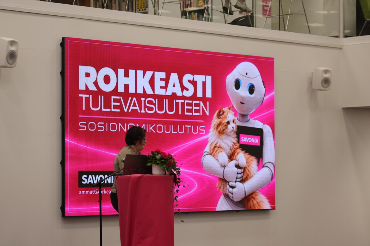 Nainen seisoo puhujakorokkeella suuren näytön edessä, jolla näkyy humanoidirobotti, joka pitää sylissään kissaa. Näytöllä lukee suomeksi: Rohkeasti Tulevaisuuteen – Sosionomikoulutus.