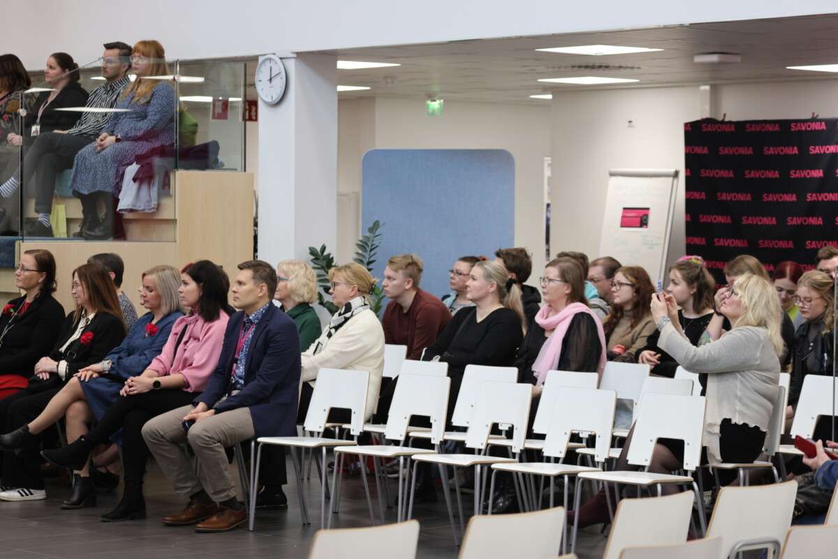 Ihmiset istuvat valkoisilla tuoleilla riveissä sisätilassa järjestettävässä tapahtumassa; osa seuraa tapahtumaa tarkkaavaisesti, osa ottaa valokuvia. Yläkerrassakin on ihmisiä, ja taustalla näkyy SAVONIA-taustakangas.