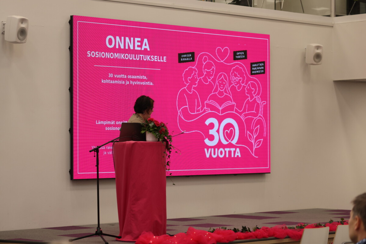 Nainen seisoo puhujakorokkeella vaaleanpunaisen taustakuvan edessä, jossa lukee ONNEA SOSIONOMIKOULUTUKSELLE 30 VUOTTA ja jossa on kuvia ihmisistä, jotka juhlivat sosiaalityön koulutuksen 30-vuotista taivalta.