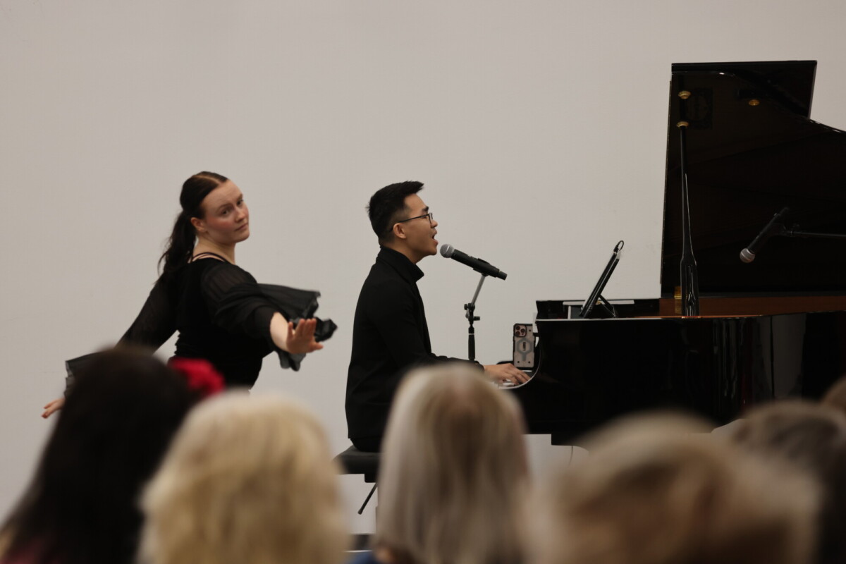 Mies soittaa pianoa ja laulaa mikrofoniin, kun taas nainen tanssii hänen vieressään. Etualalla näkyy yleisön jäsenten päitä.