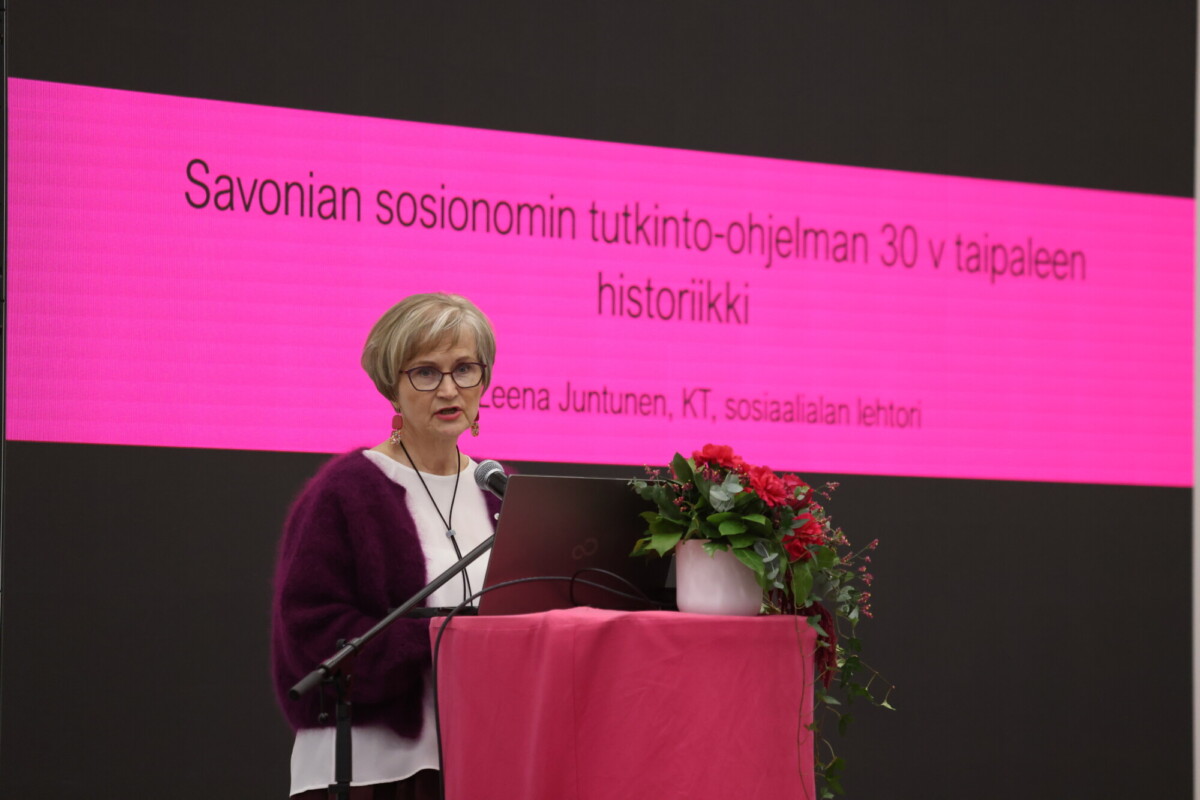 Nainen puhuu puhujakorokkeella, jonka vieressä on kannettava tietokone ja kukkia. Hänen takanaan on suuri vaaleanpunainen näyttö, jolla näkyy suomenkielistä tekstiä sosiaalityön koulutusohjelman 30-vuotisesta historiasta.