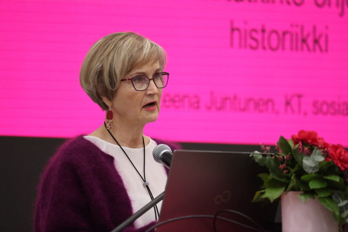 Lyhyttukkainen ja silmälasipäinen iäkkäämpi nainen puhuu puhujakorokkeella. Hänen takanaan on kirkkaan vaaleanpunainen taulu, jossa on suomalaista tekstiä. Puhujakorokkeen vieressä on punainen kukkakimppu.