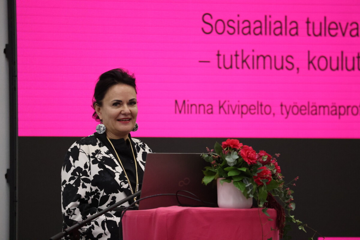 Mustavalkoiseen asuun pukeutunut nainen seisoo hymyillen kukkien kera puhujakorokkeella kirkkaan vaaleanpunaisen taustakankaan edessä, jolla näkyy suomenkielistä tekstiä.