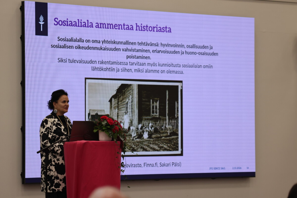 Nainen puhuu punaisella kankaalla ja kukilla koristellulla puhujakorokkeella ja esittelee dian, jonka otsikko on Sosiaaliaialla ammentaa historiasta ja jossa on tekstiä sekä vanha mustavalkoinen valokuva ihmisistä puurakennuksen lähellä.