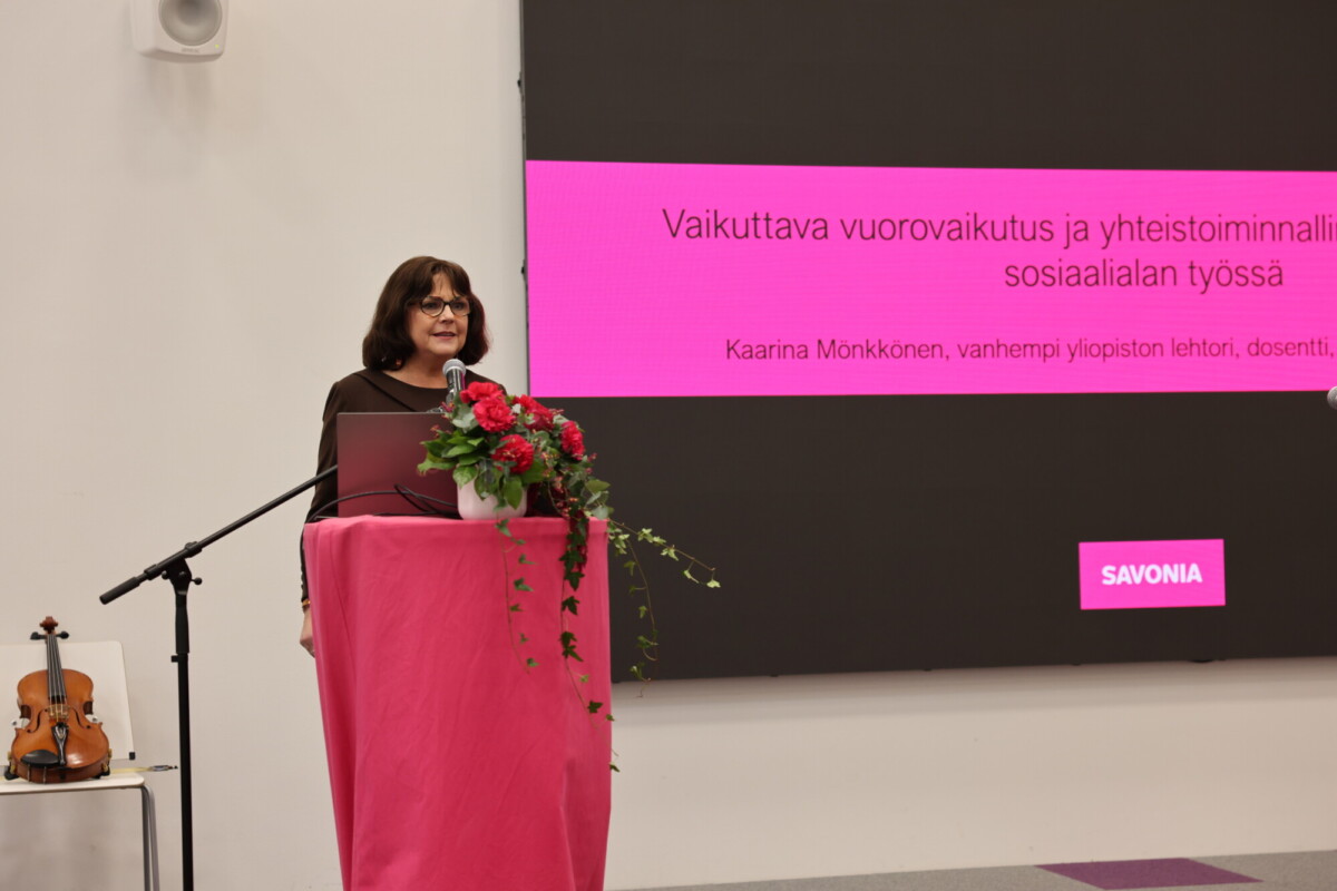Nainen seisoo puhujakorokkeella kukkakimppu kädessään ja puhuu. Hänen takanaan olevalla näytöllä näkyy tekstiä suomeksi. Viulu lepää läheisellä pöydällä. Kohtaus näyttää sijoittuvan akateemiseen tai ammatilliseen tilaisuuteen.