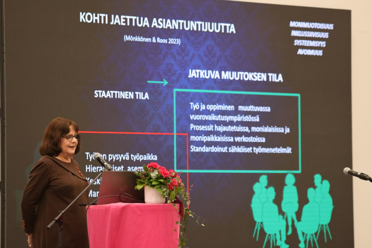 Nainen esittelee suomeksi diaesitystä, jonka aiheena on jaettu osaaminen. Diassa on värillisiä ruutuja, tekstiä ja ihmisten siluetteja. Diassa näkyy sanoja kuten monimuotoisuus ja jatkuva muutoksen tila. Puhujakorokkeella on kannettava tietokone ja kukkia.