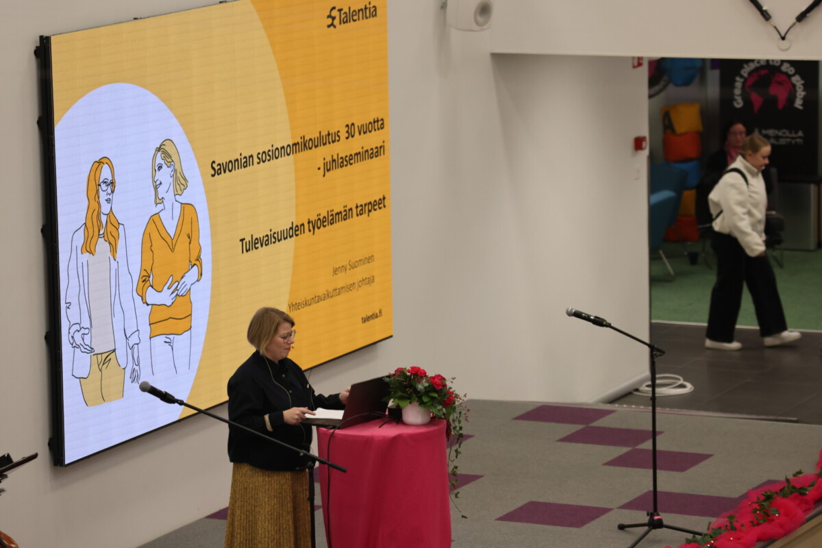 Nainen seisoo puhujakorokkeella ja puhuu suuressa salissa. Hänen takanaan esitysdia näyttää suomenkielistä tekstiä ja kuvia kahdesta naisesta. Taustalla ihmiset kävelevät.