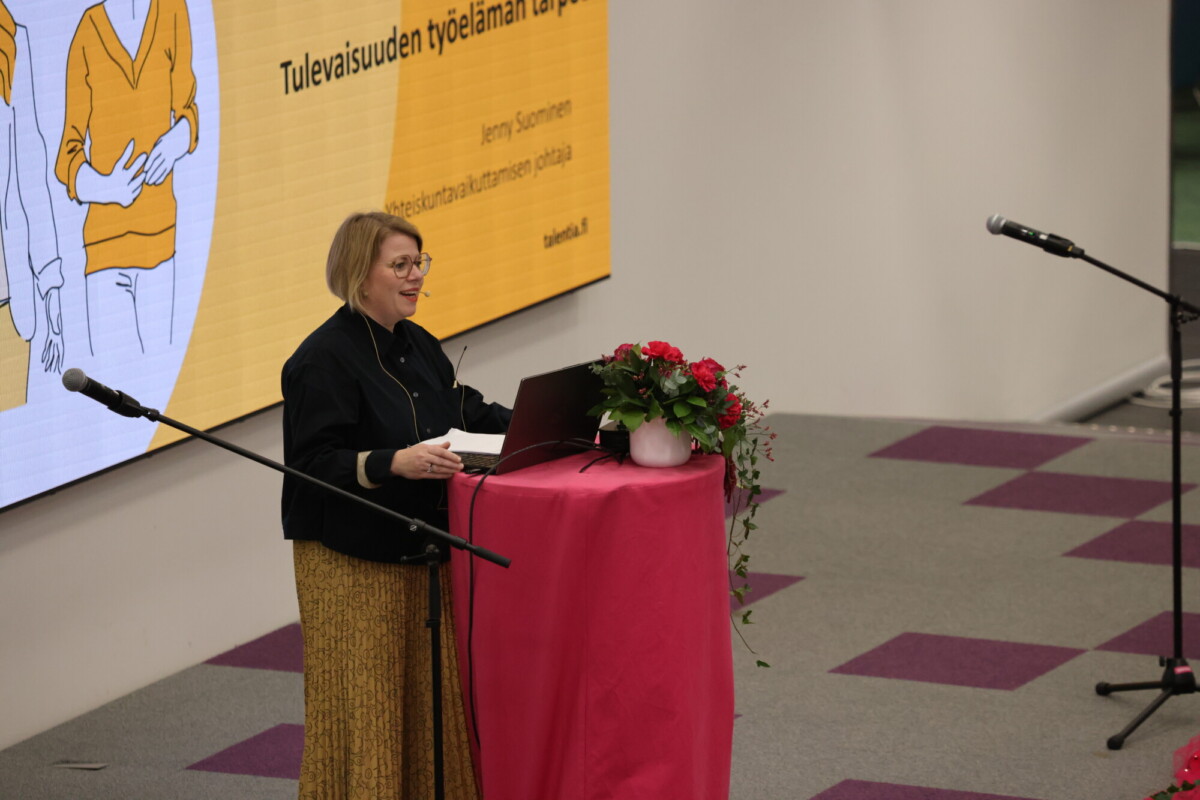 Nainen seisoo puhujakorokkeella kannettavan tietokoneen ja kukkien kanssa ja puhuu mikrofoniin. Hänen takanaan näkyy suuri keltainen esitysdia, jossa on suomenkielistä tekstiä, modernissa sisätilassa.