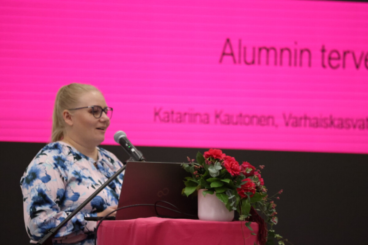 Silmälasipäinen nainen puhuu puhujakorokkeella, jonka päällä on kannettava tietokone ja kukka-asetelma. Hänen takanaan on kirkkaan vaaleanpunainen näyttö, jolla näkyy suomenkielistä tekstiä.