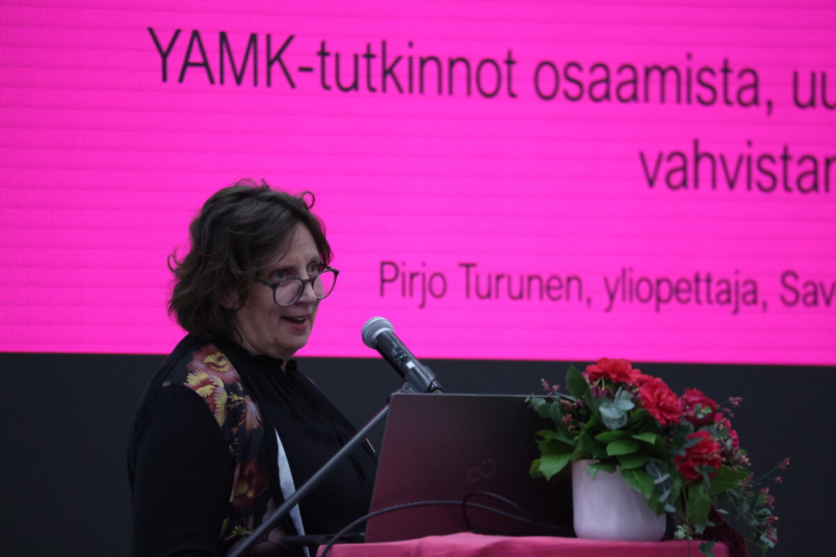 Nainen puhuu puhujakorokkeella, jonka päällä on kannettava tietokone ja kukkia, kirkkaan vaaleanpunaisen näytön edessä, jolla näkyy suomenkielistä tekstiä koulutuksesta sekä nimi Pirjo Turunen.