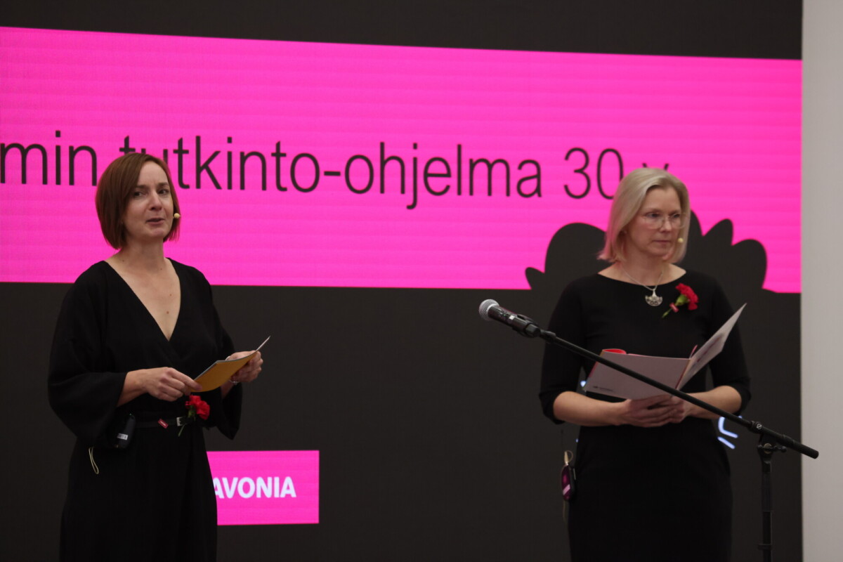 Kaksi mustissa mekkoissa olevaa naista seisoo lavalla papereita kädessään, ja lähellä on mikrofoni. Heidän takanaan olevalla suurella näytöllä näkyy suomenkielistä tekstiä mustalla ja vaaleanpunaisella taustalla.