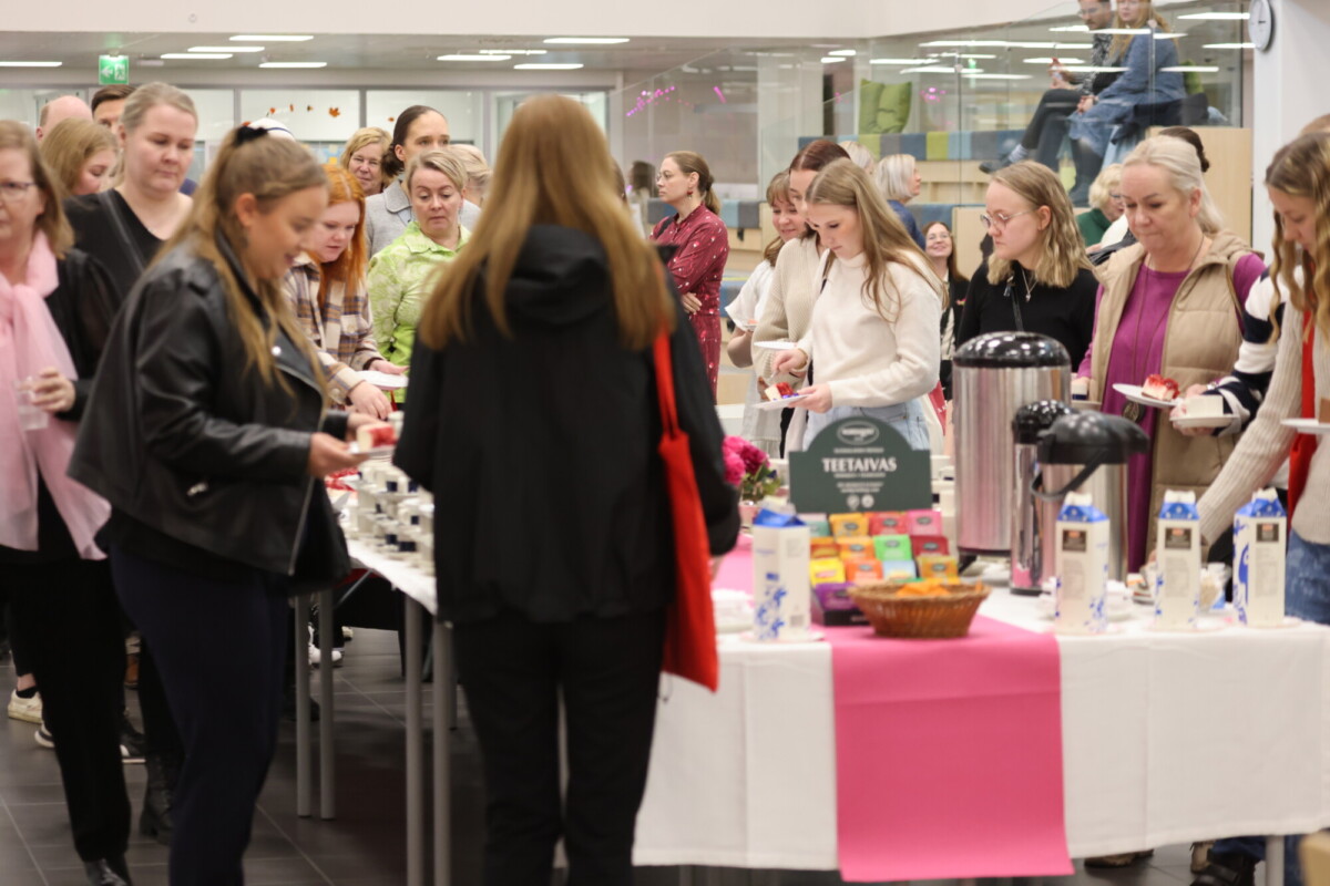 Ryhmä ihmisiä on kokoontunut pöydän ympärille, jolla on kahvia, maitopakkauksia, kuppeja ja välipaloja, valoisassa sisätilassa. Osa ottaa itse ruokaa, kun taas toiset odottavat jonossa.
