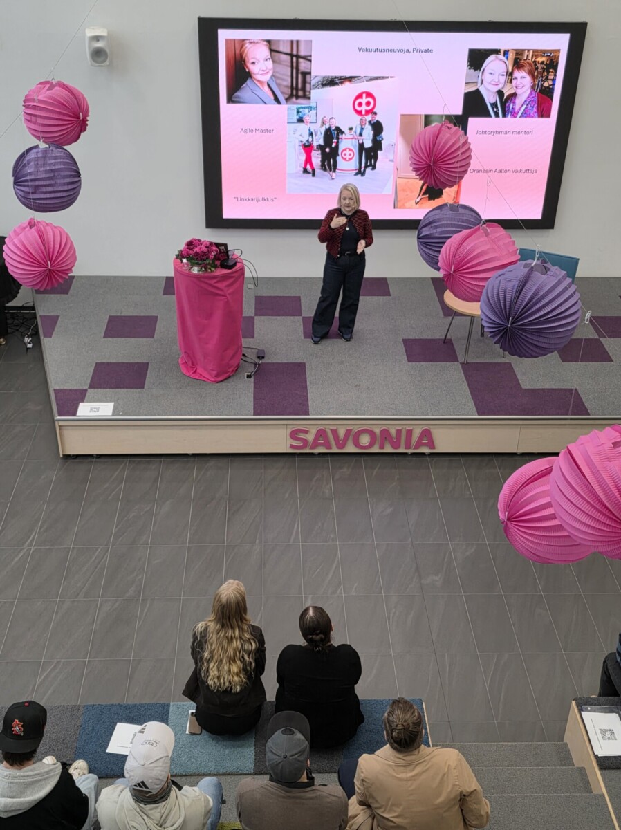 Nainen puhuu lavalla, jonka edessä on suuri näyttö, jolla näkyy valokuvia ja tekstiä, kun taas yleisö seuraa esitystä alhaalta, jossa tilaa koristavat vaaleanpunaiset ja violetit paperilyhdyt.