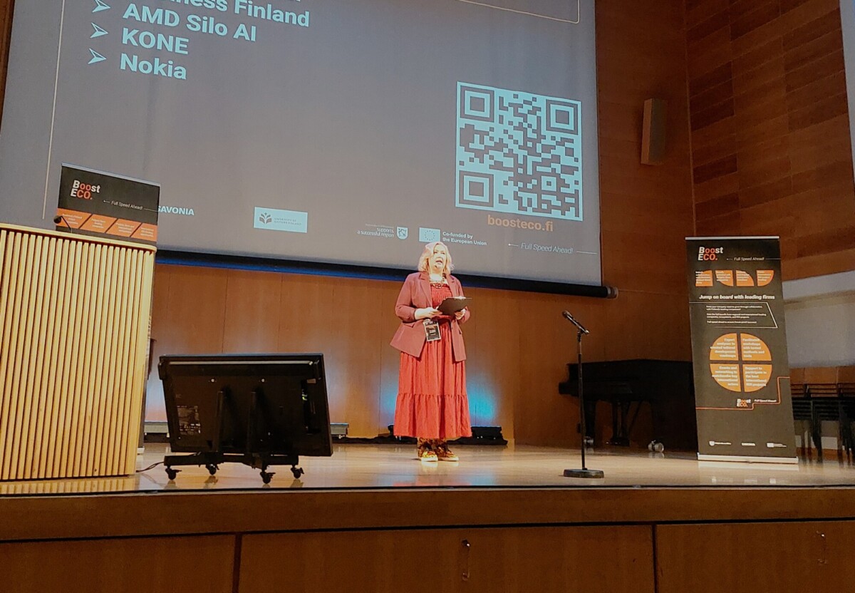 Punaisessa mekossa oleva nainen seisoo lavalla ja puhuu puhujakorokkeelta projektorinäytön edessä, jolla näkyy QR-koodi ja yritysten luettelo.