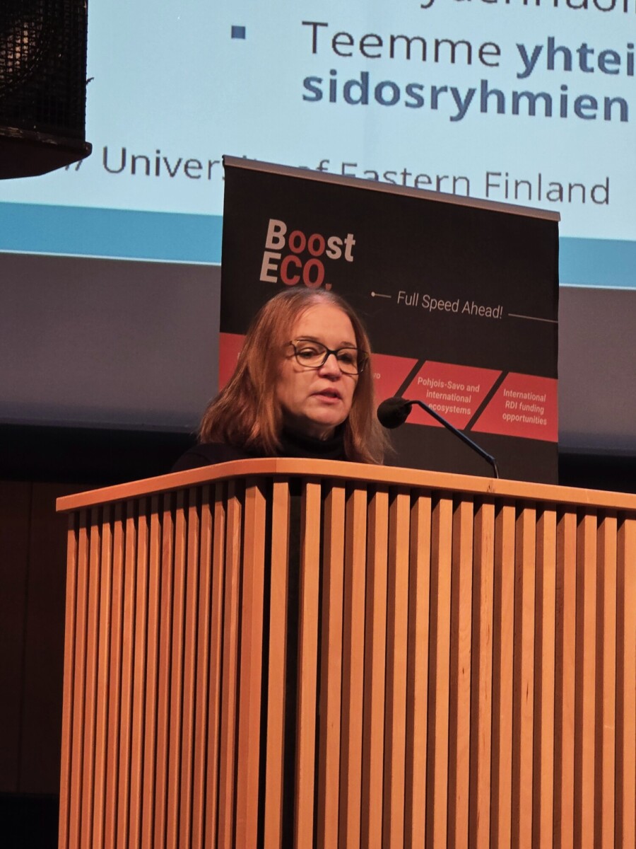 Silmälasipäinen nainen puhuu puisen puhujakorokkeen ääressä esitysnäytön ja Boost ECO -merkkisen banderollin edessä.