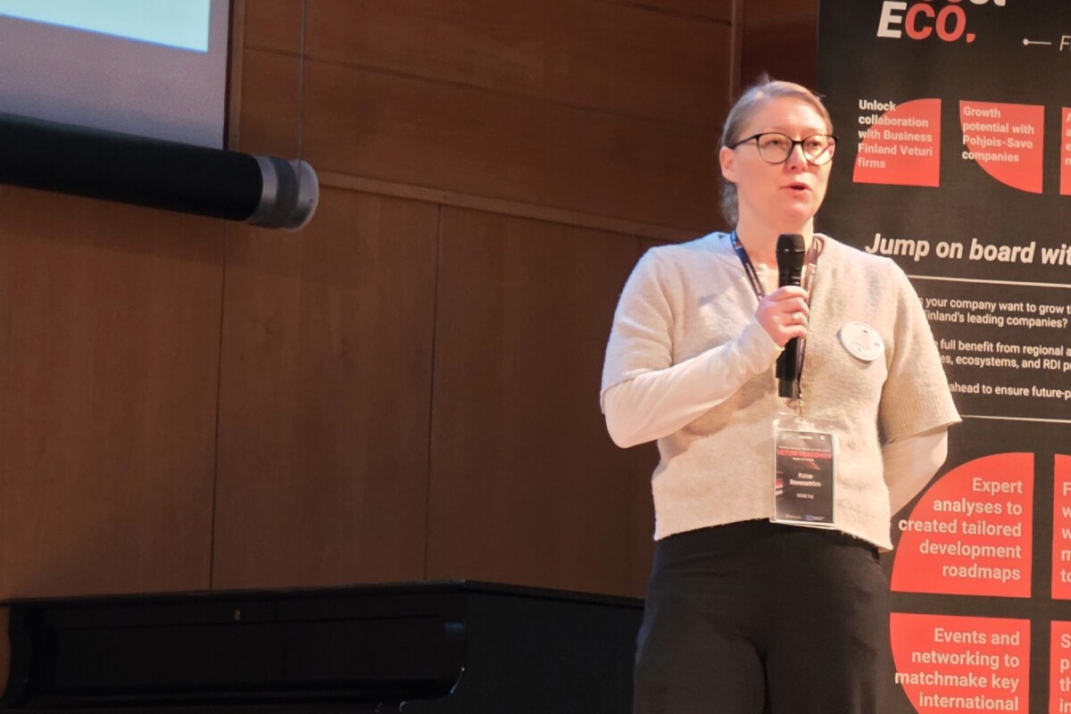 Lasit päässä oleva nainen puhuu mikrofoniin esityksessä, jonka taustalla on liiketoimintayhteistyötä kuvaava kyltti.