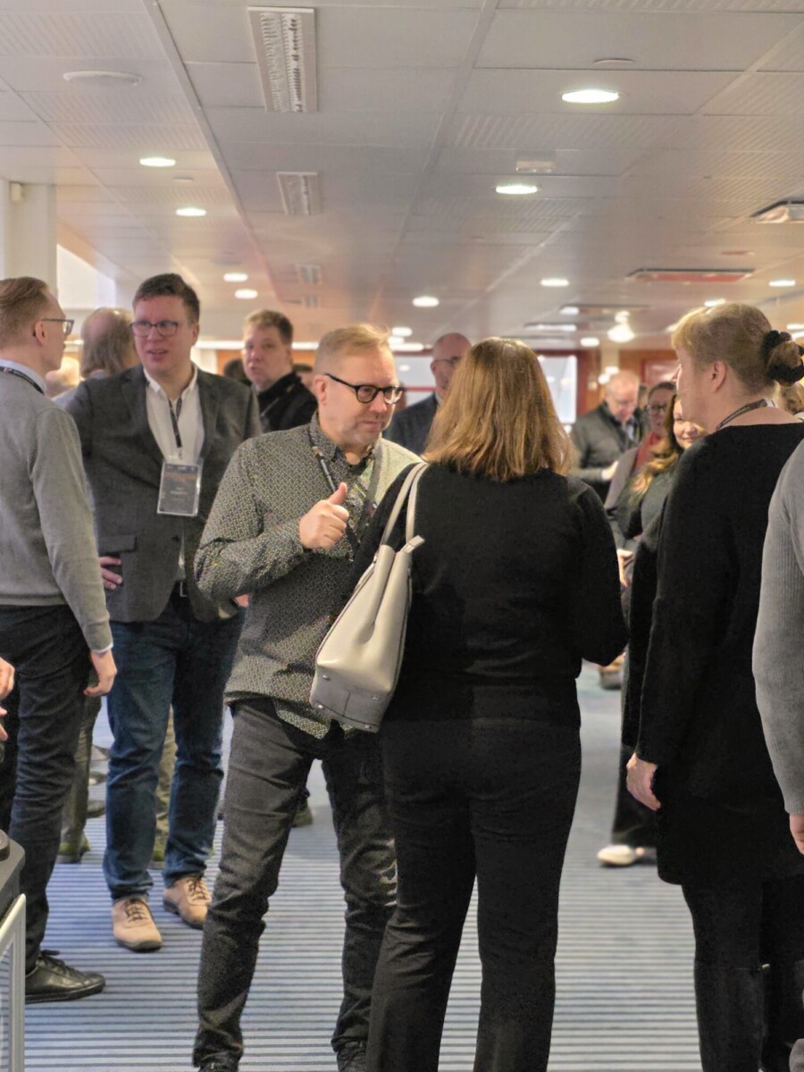 Mies, jolla on kuviollinen napituspaita, näyttää peukaloa ylöspäin puhuessaan mustassa villapaidassa olevalle naiselle ruuhkaisessa käytävässä.