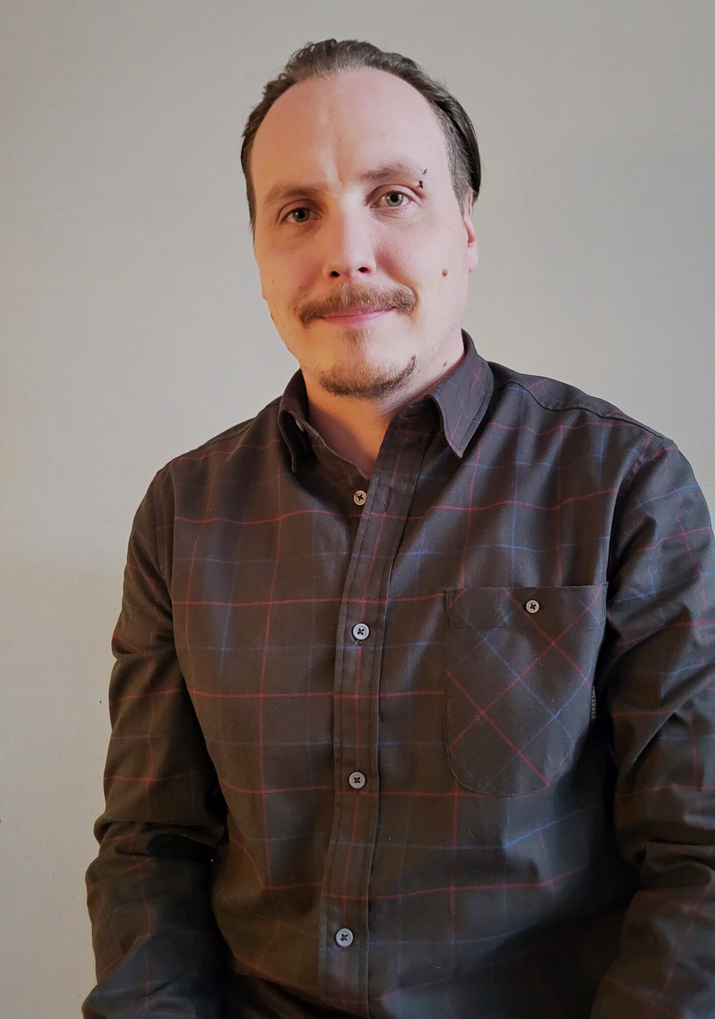 Mies, jolla on viikset ja lävistys vasemman kulmakarvan yläpuolella ja joka on pukeutunut tummaan ruudulliseen napituspaitaan.