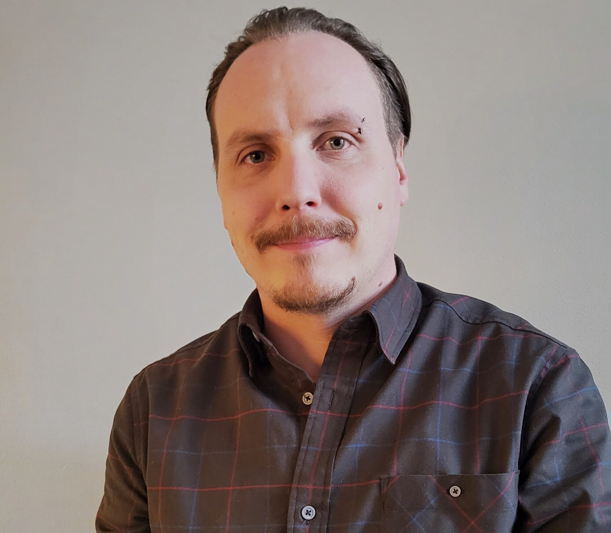 Mies, jolla on viikset ja lävistys kulmakarvan yläpuolella ja joka on pukeutunut tummaan ruudulliseen napituspaitaan.