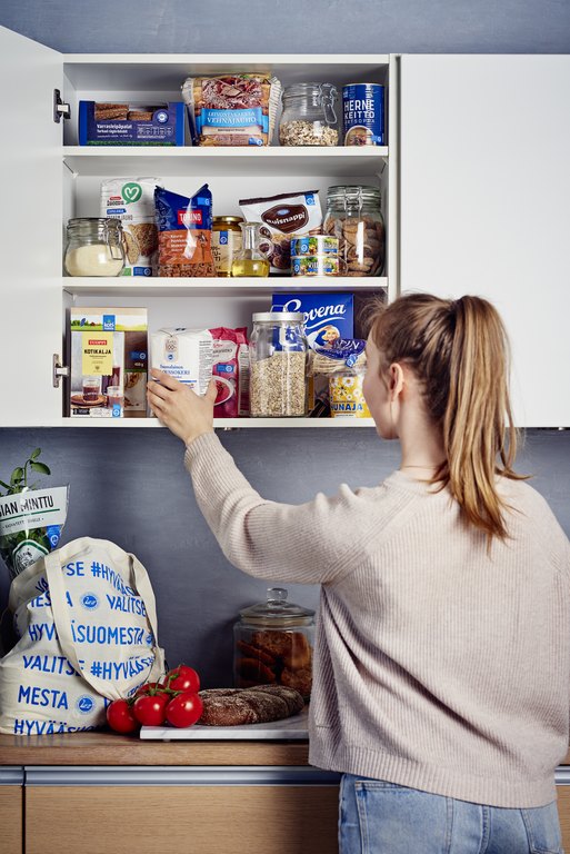 Henkilö kurkottaa avoimeen keittiökaappiin, joka on täynnä erilaisia elintarvikkeita pakkauksissa ja purkeissa.