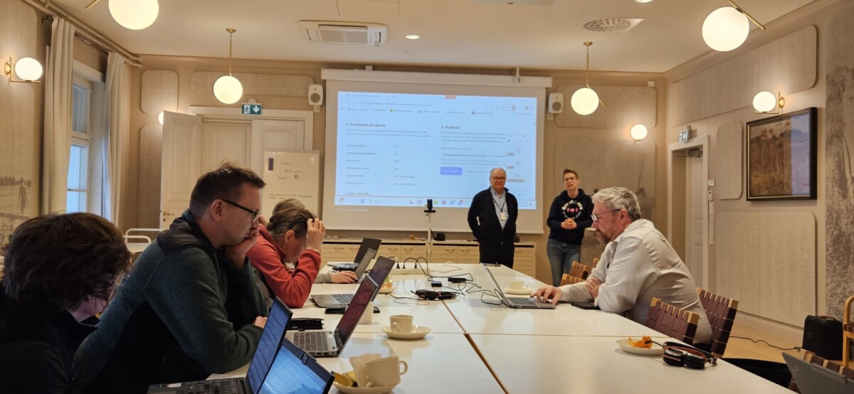 Ryhmä ihmisiä istuu pitkän kokouspöydän ympärillä ja katselee kannettavia tietokoneita, kun taas yksi mies seisoo ja pitää esitystä valkokankaan edessä, jolla näkyy verkkosivusto.