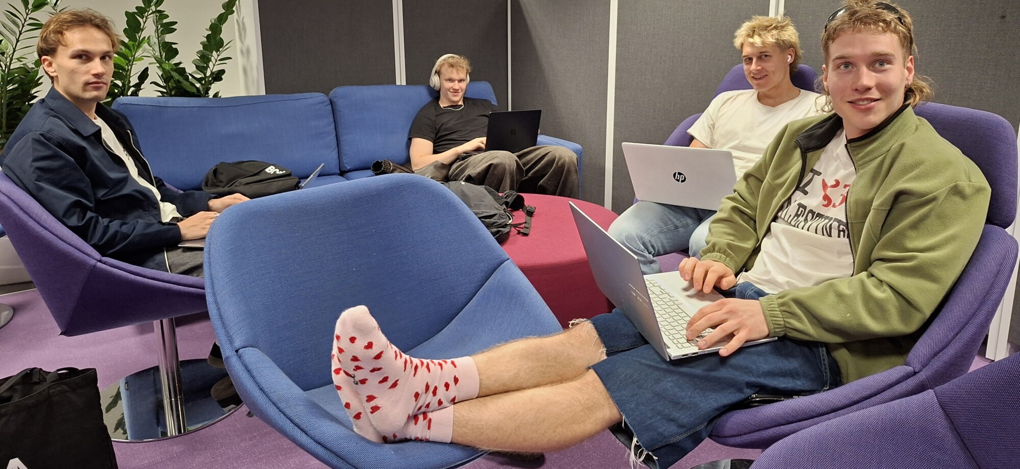Neljä nuorta miestä istuu oleskelutilassa kannettavia tietokoneita käyttäen, ja etualalla on yksi henkilö, jolla on sydämenkuvioiset sukat.