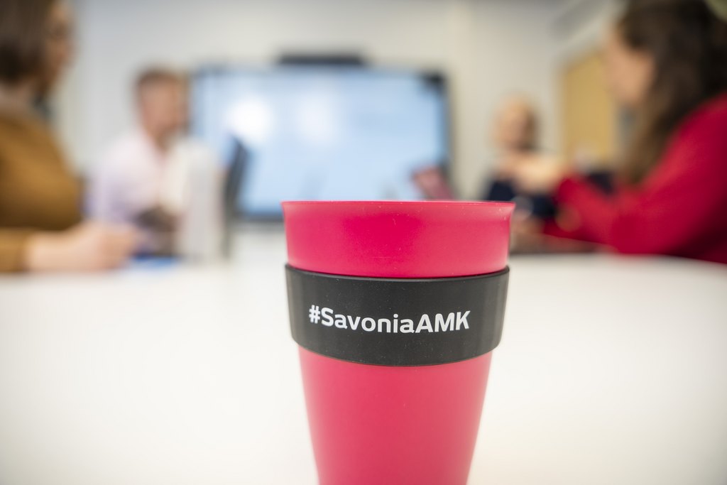 Hämärtyneen toimistokokouksen etualalla näkyy vaaleanpunainen uudelleenkäytettävä kuppi, jonka mustassa suojuksessa lukee #SavoniaAMK.