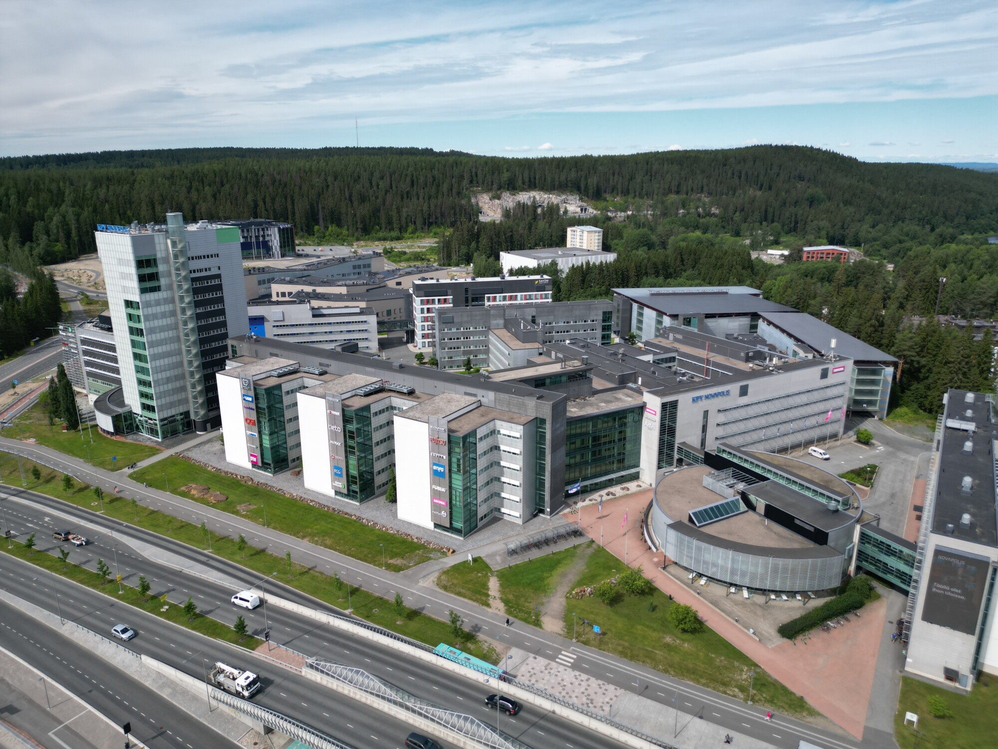 Ilmakuva modernista yliopistokampuksesta, jossa on useita suuria rakennuksia ja jota ympäröivät tiet, autot ja vihreät metsäalueet osittain pilvisen taivaan alla.