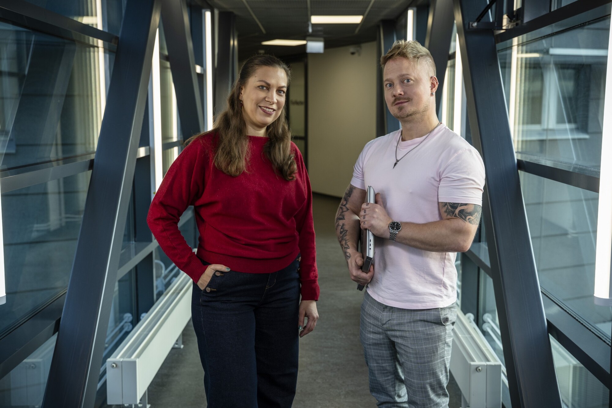 Punaisessa villapaidassa oleva nainen ja vaaleanpunaisessa t-paidassa oleva mies, joka pitää sylissään kannettavaa tietokonetta, seisovat modernissa, lasiseinäisessä käytävässä.