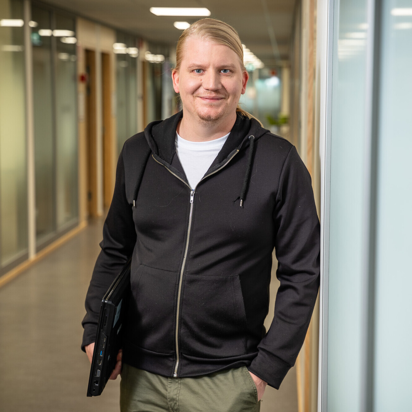 Vaaleatukkainen mies, jolla on yllään musta vetoketjullinen huppari ja khakinväriset housut, seisoo käytävällä suljettua kannettavaa tietokonetta kädessään ja hymyilee kameraan. Käytävällä on lasiseinäisiä toimistoja ja kirkas valaistus.