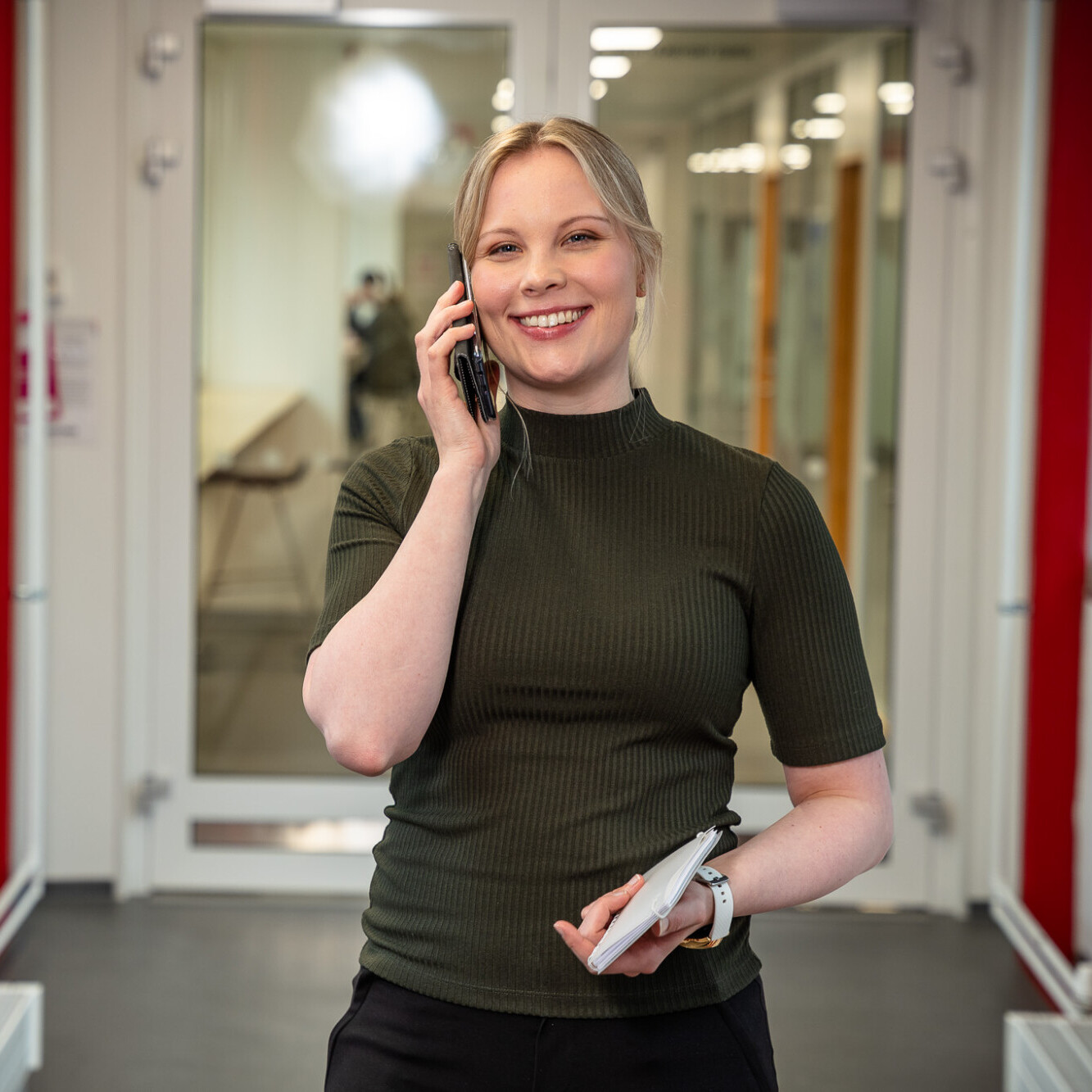 Nainen seisoo punaisilla palkkeilla koristellussa käytävässä, hymyillen puhuessaan puhelimeen ja pitäen toisessa kädessään muistikirjaa ja kynää.
