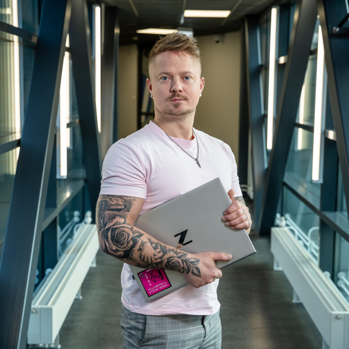 Tatuoitu henkilö, jolla on vaaleanpunainen paita, seisoo modernissa lasisessa käytävässä, jossa on mustia metallipalkkeja, ja pitää kädessään kannettavaa tietokonetta ja nimikylttiä.