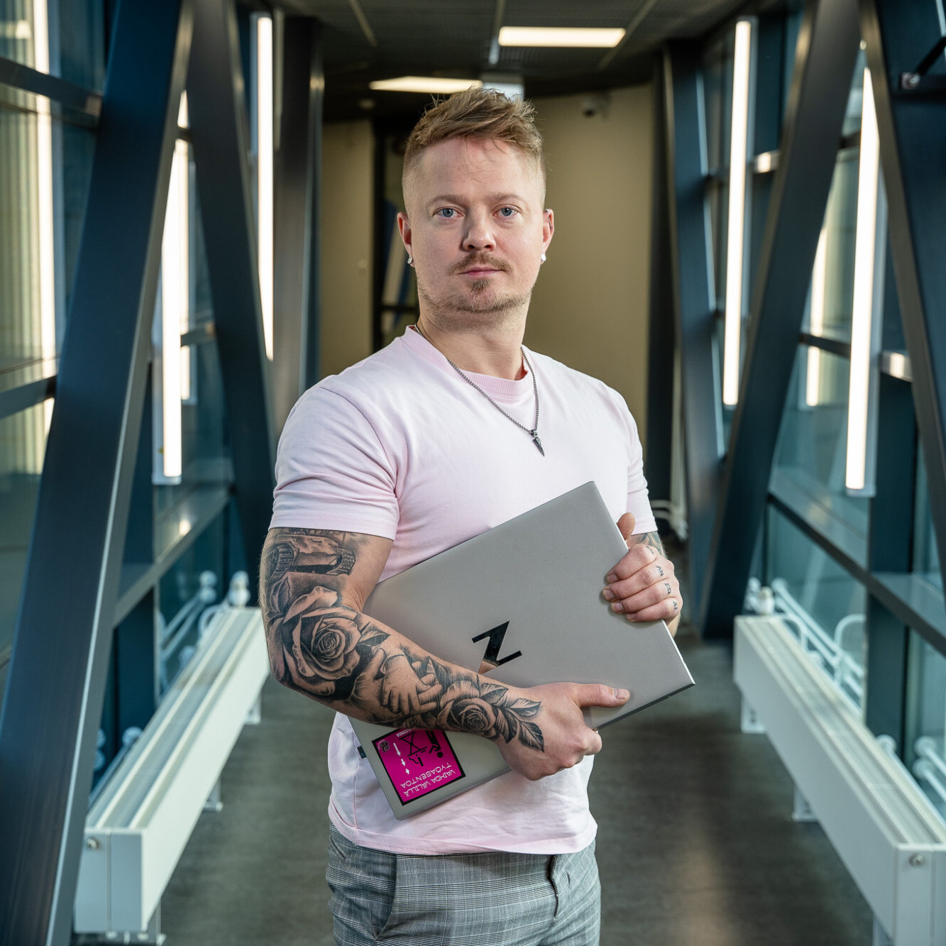 Tatuoitu henkilö, jolla on vaaleanpunainen paita, seisoo modernissa lasisessa käytävässä, jossa on mustia metallipalkkeja, ja pitää kädessään kannettavaa tietokonetta ja nimikylttiä.