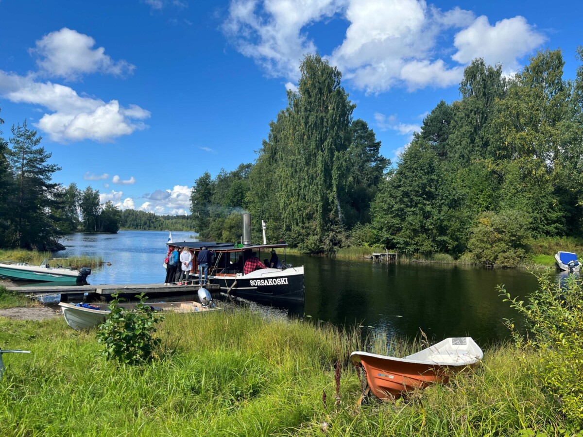 Ihmiset nousevat pienelle Sorsakoski-nimiselle veneelle, joka on kiinnitetty ruohikkoiseen rantapenkkiin järven rannalla.