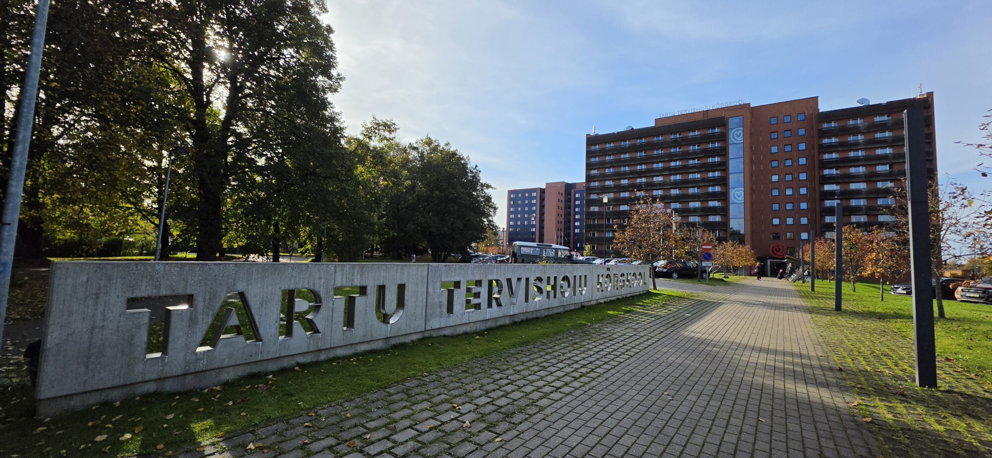 Aurinkoisena päivänä monikerroksisen rakennuksen edessä on betonikyltti, jossa lukee TARTU TERVISHOIU KÕRGKOOL.