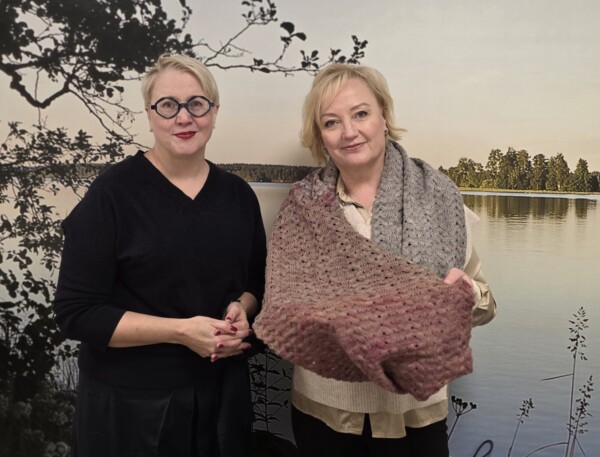 Kaksi naista seisoo järvenrannan seinämaalauksen edessä, ja toinen heistä pitää kädessään kuvioitua, moniväristä neulottua huivia.