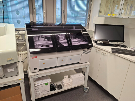 Valkoinen laboratoriomittari seisoo metallisella vaunulla huoneessa, jonka läheisyydessä on tietokoneen näyttö ja näppäimistö.