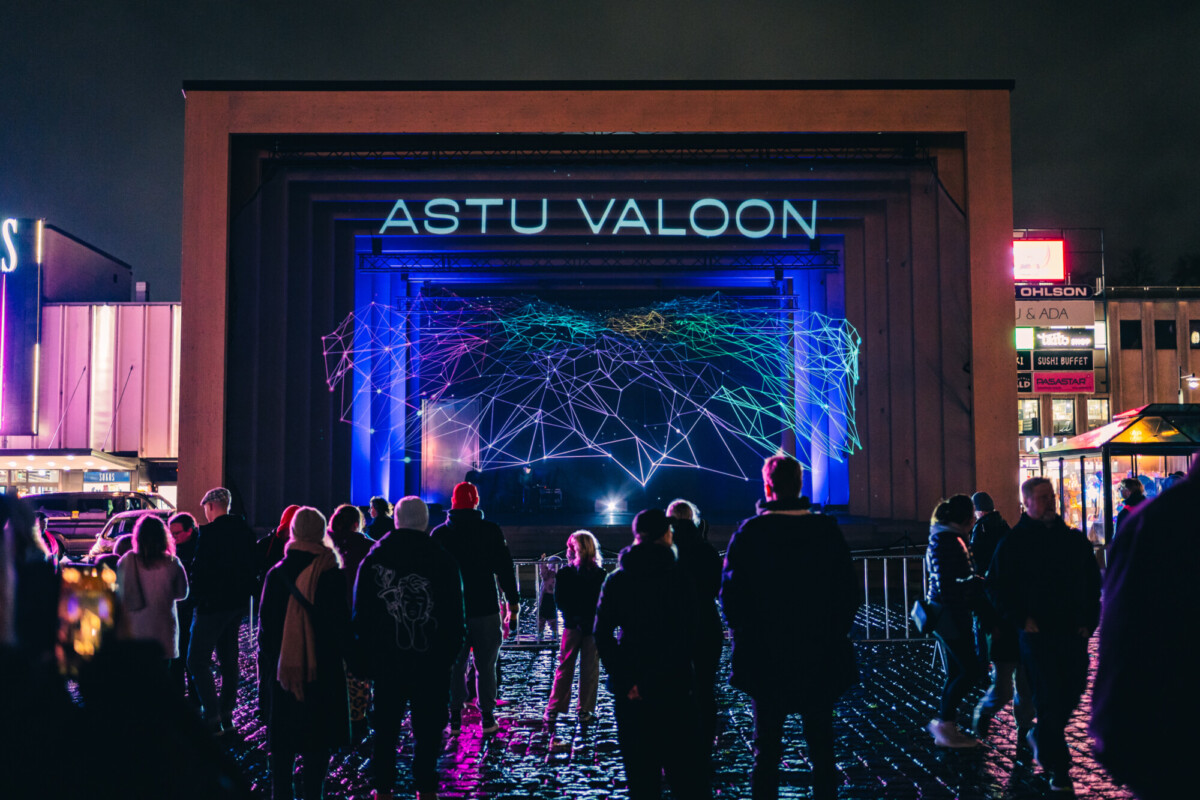 Ihmisjoukko seisoo lavan edessä, jolla on esillä hohtava, geometrinen verkkomainen taideinstallaatio sekä sanat ASTU VALOON neoninsinisillä kirjaimilla.