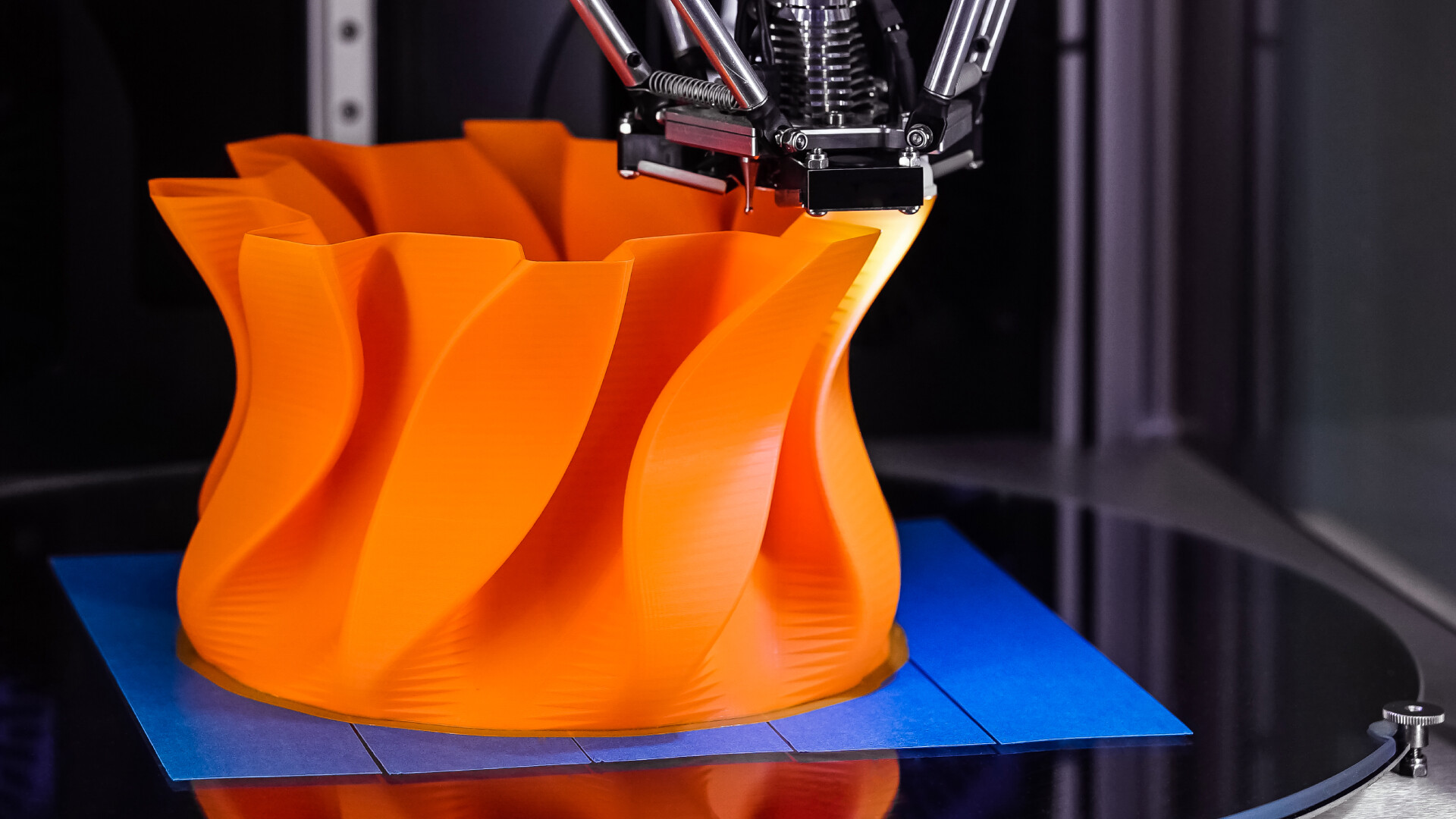 3D-tulostimen suutin levittää oranssia filamenttia luodakseen kierteisen maljakon muodon tulostusalustalle.