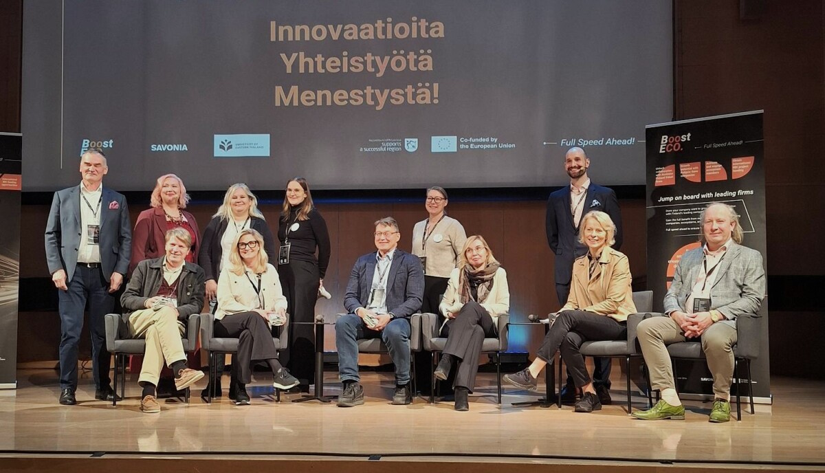 Yksitoista hengen ryhmä, joka seisoo ja istuu lavalla esitysnäytön ja projektibannerin edessä.