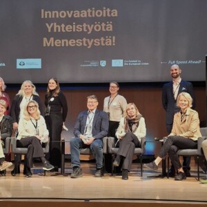 Yksitoista hengen ryhmä, joka seisoo ja istuu lavalla esitysnäytön ja projektibannerin edessä.
